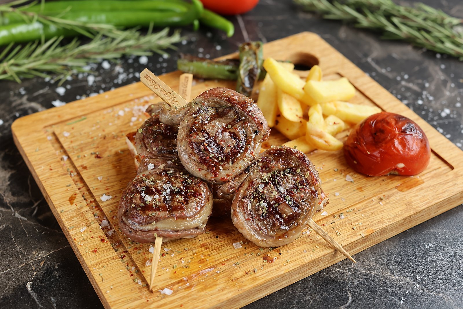 Grilled lamb medallions on skewers served on a wooden board with fries, roasted tomato and green chilli