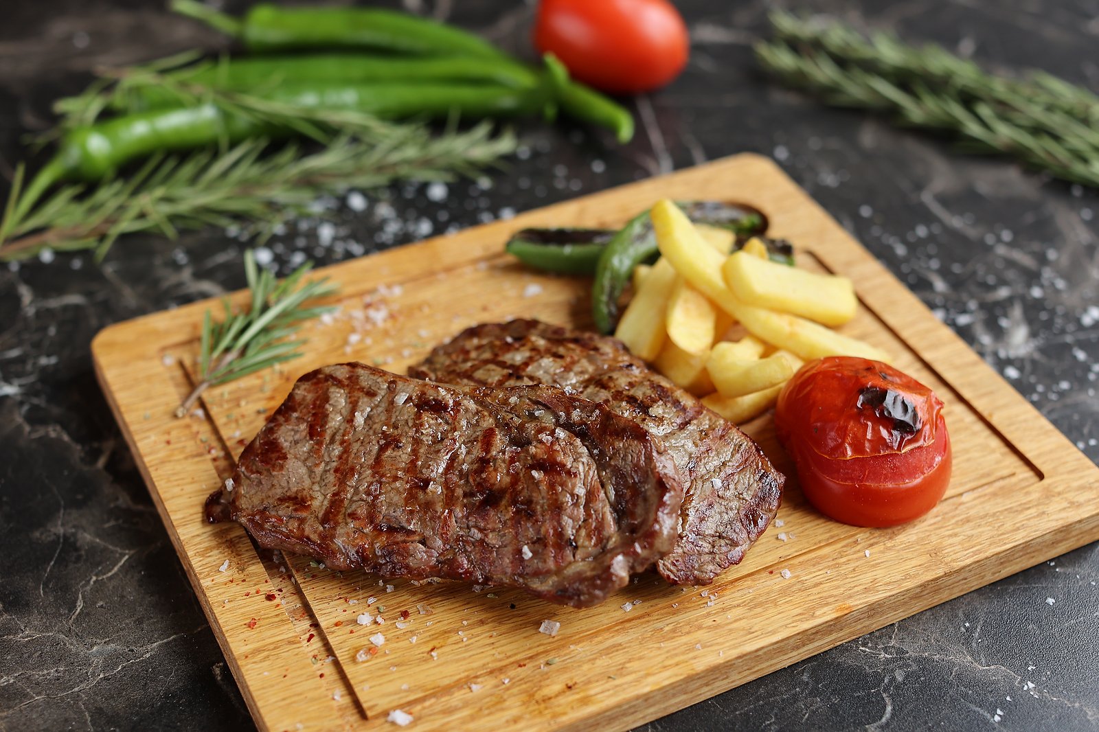 Grilled steak on a wooden board with fries, roasted tomato and green chillies